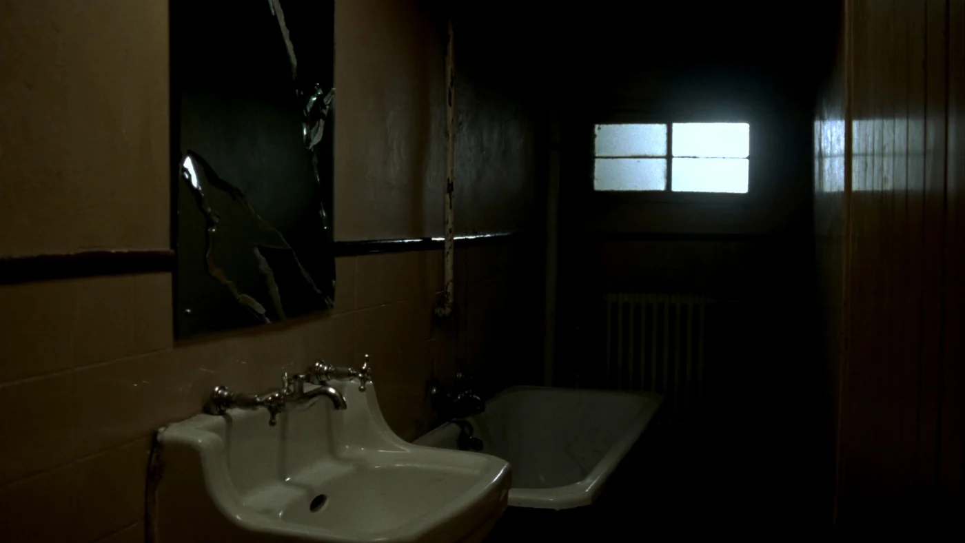 Salle de bains sombre avec un miroir fissuré à gauche, un lavabo au premier plan et une fenêtre claire au fond.
