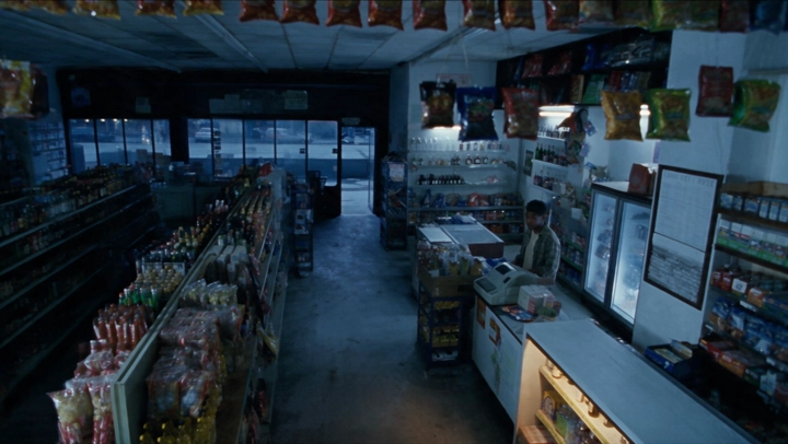 Vue en plongée d’une épicerie : allées en diagonale, réfrigérateurs au fond et Marcus au comptoir à droite.
